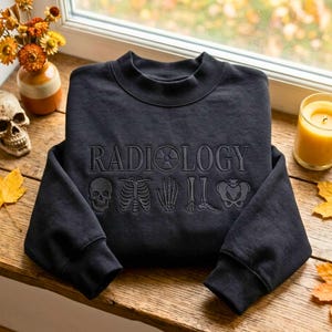 May include: A black crewneck sweatshirt featuring the word RADIOLOGY embroidered in a dark thread across the chest. The letter O is replaced by a radiation symbol. Below the text are embroidered outlines of a skull, ribcage, hand, foot, and pelvis.
