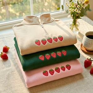 May include: Three folded sweatshirts in beige, green, and pink, stacked on top of each other. Each sweatshirt has a row of embroidered strawberries. The beige sweatshirt has a zipper at the collar. Fresh strawberries and a cup of coffee are nearby.