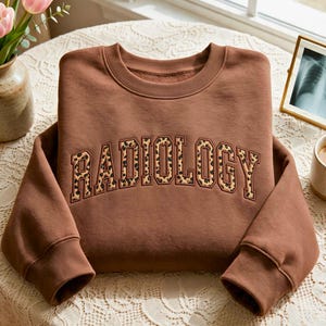 以下が含まれることがあります： 胸元にカレッジスタイルのフォントで「RADIOLOGY」という文字が刺繍されたブラウンのクルーネックトレーナー。文字はブラウンとブラックのヒョウ柄で埋め尽くされており、ダークブラウンの糸で縁取りが施され、立体感のある仕上がりになっています。