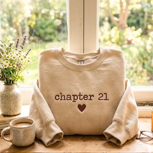 Puede incluir: Una sudadera beige de cuello redondo con el texto "chapter 21" en marrón y un corazón marrón. La sudadera está doblada sobre una superficie de madera, con una taza de café, un jarrón de flores y un par de gafas en el fondo.