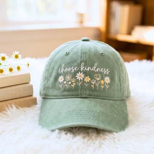 Könnte beinhalten: Eine salbeigrüne Baseballkappe mit dem gestickten weißen Schriftzug choose kindness über einer Reihe von weißen und gelben Wildblumen. Die Kappe hat eine strukturierte Krone, einen gebogenen Schirm und Ösen zur Belüftung an den Paneelen.