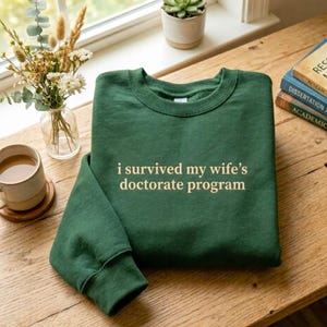May include: A dark green sweatshirt with the text "i survived my wife's doctorate program" in a white, sans-serif font. The sweatshirt is folded on a wooden surface, with a coffee mug, flowers, and books in the background.