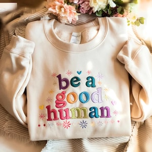May include: Cream-coloured sweatshirt embroidered with the phrase "be a good human" in colourful letters. The design includes embroidered stars and hearts. The sweatshirt is folded on a textured surface, with flowers in the background.
