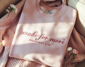 Könnte beinhalten: Hellrosa Sweatshirt mit der gestickten Aufschrift "made for more" und dem Text "Ephesians 2:10" in Schreibschrift. Das Sweatshirt liegt gefaltet auf einer rosa Decke, mit neutralem Hintergrund und floralen Akzenten.