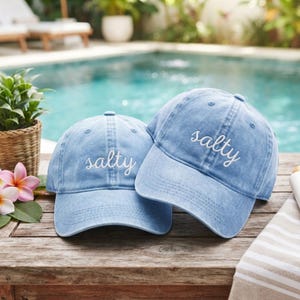 May include: Two light blue denim baseball caps with a distressed finish, each featuring the word "salty" embroidered in white cursive script on the front panel. The caps have a structured crown, a curved brim, and matching stitching details.