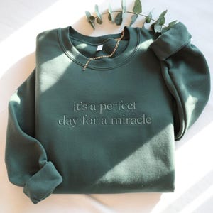 May include: Dark green sweatshirt with the embroidered text "it's a perfect day for a miracle." The long-sleeved top is folded neatly, with a gold chain draped across the neckline. Eucalyptus leaves are in the background.