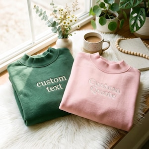 May include: A dark green sweatshirt featuring the embroidered text custom text and a light pink sweatshirt with the embroidered text Custom Quote. Both garments have a mock neck collar, long sleeves, and are made from a soft, solid-coloured fabric.