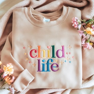 Könnte beinhalten: Beiges Sweatshirt mit dem gestickten Text "child life" in Regenbogenfarben. Das Design umfasst Herzen und Sterne. Das Sweatshirt liegt gefaltet auf einer rosa Decke mit Blumen.