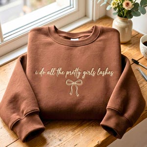 以下が含まれることがあります： 胸元に「i do all the pretty girls lashes」という明るい色の筆記体テキストと、その下にシンプルな線画のリボンのイラストが描かれた、茶色のクルーネックのスウェットシャツ。
