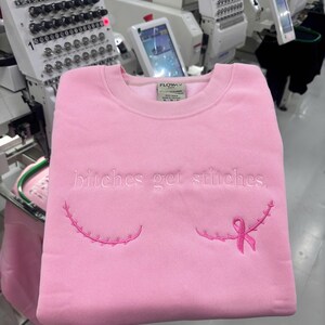Pode incluir: Uma camisola de gola redonda rosa claro com o texto bordado "bitches get stitches." em fio rosa claro. Abaixo do texto, existem duas linhas curvas bordadas que lembram cicatrizes cir&uacute;rgicas, com um la&ccedil;o rosa de consciencializa&ccedil;&atilde;o &agrave; direita.