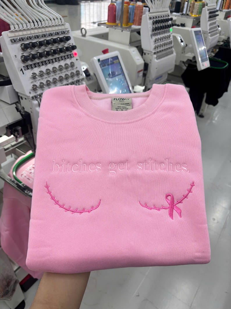 Pode incluir: Uma camisola de gola redonda rosa claro com o texto bordado "bitches get stitches." em fio rosa claro. Abaixo do texto, existem duas linhas curvas bordadas que lembram cicatrizes cir&uacute;rgicas, com um la&ccedil;o rosa de consciencializa&ccedil;&atilde;o &agrave; direita.