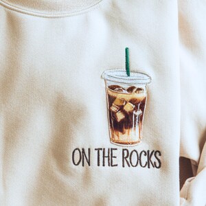 May include: Cream-colored sweatshirt featuring an embroidered iced coffee design with a green straw. The phrase "ON THE ROCKS" is embroidered below the coffee cup. A casual, comfortable top for coffee lovers.