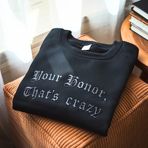 May include: Black sweatshirt with the embroidered phrase "Your Honor, That's crazy" in a gothic font. The sweatshirt is folded on a brown cushion, with a wooden side table and books in the background.