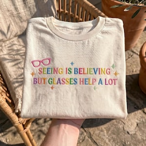 May include: A cream-coloured t-shirt featuring embroidered text that reads "SEEING IS BELIEVING BUT GLASSES HELP A LOT" in multicoloured letters. Above the text is an embroidered pink pair of glasses, surrounded by small embroidered stars.