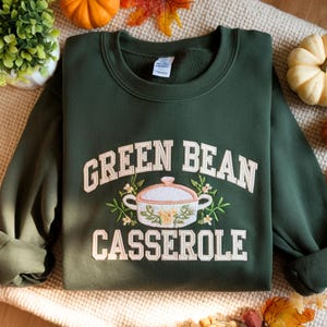 May include: Dark green sweatshirt with the words "GREEN BEAN CASSEROLE" in white embroidered lettering. A casserole dish with floral accents is centered below the text. The sweatshirt is displayed on a neutral surface with fall-themed decorations.
