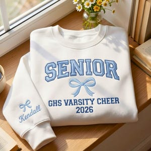 May include: A white sweatshirt with light blue embroidered text and bow details. The word "SENIOR" is across the chest, with "GHS VARSITY CHEER 2026" below. The sleeve has a bow and the name "Kendall".