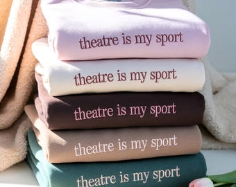 May include: A stack of five crewneck sweatshirts in light pink, cream, dark brown, tan, and dark green. Each sweatshirt features the embroidered text "theatre is my sport" in a contrasting colour centred on the chest area of the folded garment.