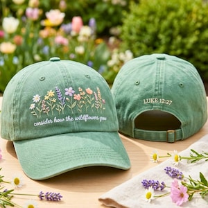 May include: A sage green baseball cap featuring embroidered colorful wildflowers and the white text "consider how the wildflowers grew" on the front, with "LUKE 12:27" embroidered in white above the adjustable back strap.