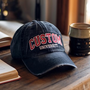 May include: A distressed navy blue baseball cap with the words "CUSTOM UNIVERSITY" embroidered in red and white. The cap sits on a wooden table next to an open book and a ceramic mug.