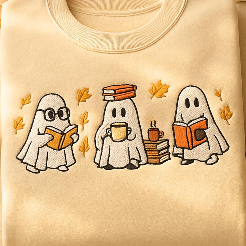 Puede incluir: Sudadera de color crema con fantasmas de dibujos animados bordados. Un fantasma lleva gafas y lee un libro, otro sostiene una taza y el tercero lee un libro. Hojas de oto&ntilde;o est&aacute;n esparcidas alrededor de los fantasmas.