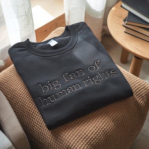 Puede incluir: Sudadera negra de cuello redondo con la frase bordada "big fan of human rights". La sudadera est&aacute; doblada y colocada sobre un coj&iacute;n tejido. El texto est&aacute; en una fuente sencilla, sin serifa.