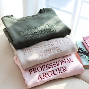 May include: Three sweatshirts stacked. The top one is olive green with embroidered text: "PROFESSIONAL LISTENER". The middle one is light beige with "PROFESSIONAL YAPPER". The bottom one is pink with "PROFESSIONAL ARGUER". The sweatshirts are on a light-colored surface.