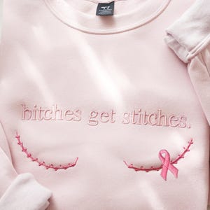 Pode incluir: Uma camisola de gola redonda rosa claro com o texto bordado "bitches get stitches." em fio rosa a condizer. Abaixo do texto, existem duas linhas curvas de bordado rosa que se assemelham a pontos cir&uacute;rgicos, com um la&ccedil;o rosa &agrave; direita.