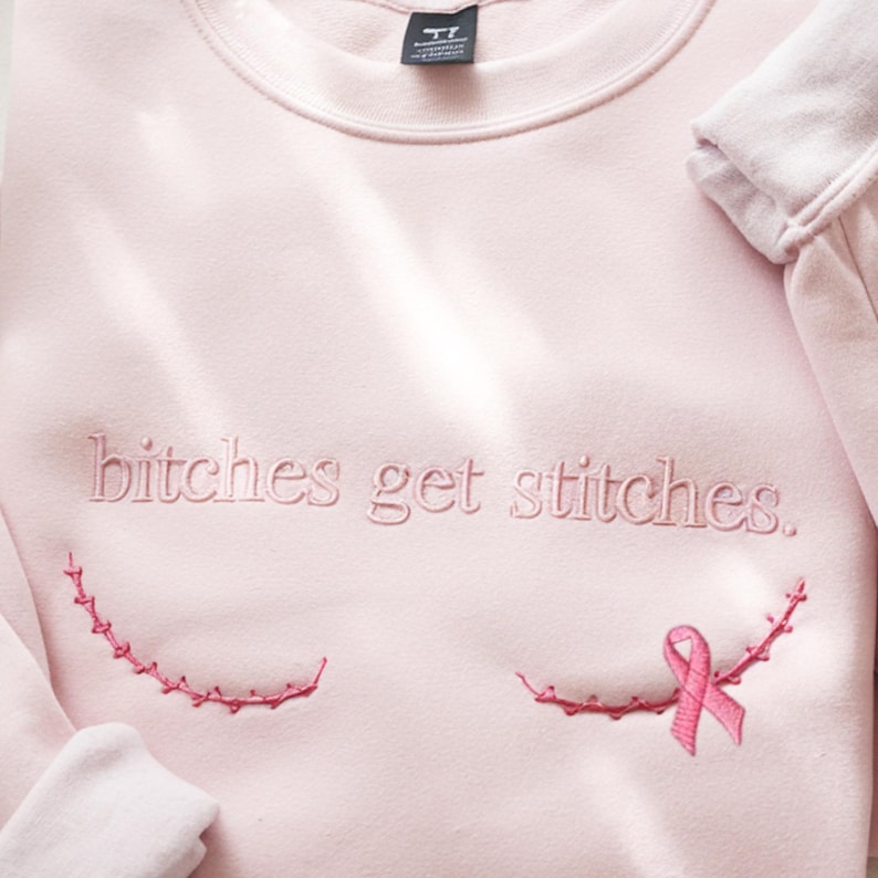 Pode incluir: Uma camisola de gola redonda rosa claro com o texto bordado "bitches get stitches." em fio rosa a condizer. Abaixo do texto, existem duas linhas curvas de bordado rosa que se assemelham a pontos cir&uacute;rgicos, com um la&ccedil;o rosa &agrave; direita.