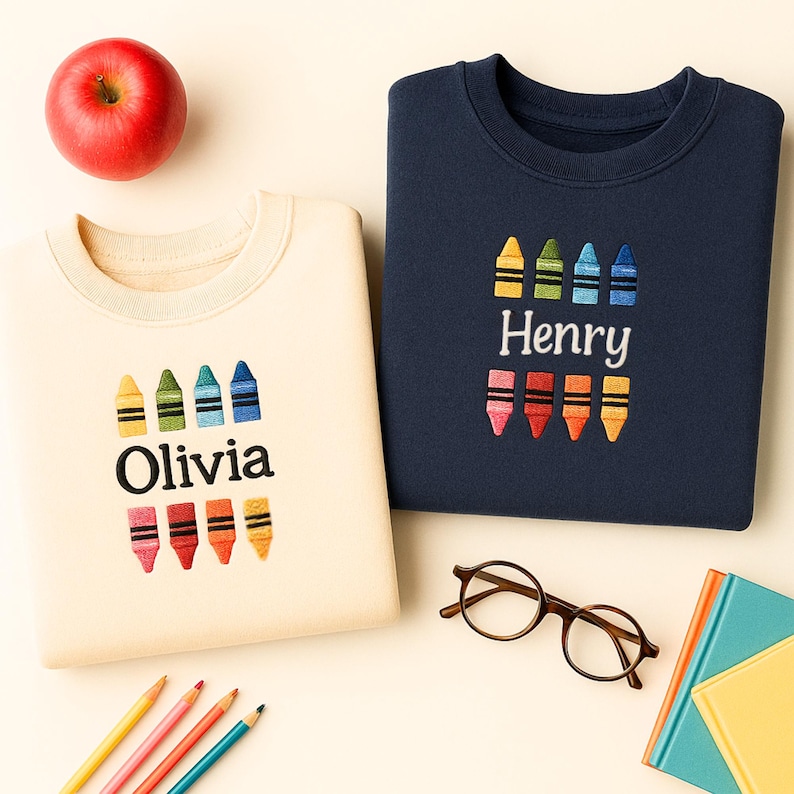 Op de afbeelding: Twee sweatshirts, &eacute;&eacute;n cr&egrave;me en &eacute;&eacute;n marineblauw, elk geborduurd met kleurrijke kleurpotloden en een naam. Het cr&egrave;mekleurige sweatshirt zegt "Olivia", en de marineblauwe zegt "Henry". Een rode appel, potloden, een bril en een notitieboek staan ook in de afbeelding.