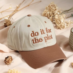 May include: A beige baseball cap with a brown brim and the embroidered phrase "do it for the plot" in brown thread. The cap has a curved brim and a button on top. The hat is a casual accessory.