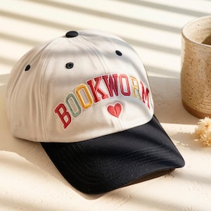 May include: A baseball cap with a cream-colored crown and a black brim. The word "BOOKWORM" is embroidered in colorful letters across the front, with a red heart below. A black button is on top.