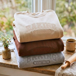 May include: A stack of three crewneck sweatshirts in beige, brown, and heather gray. Each sweatshirt features the embroidered text BAKING WEATHER in a collegiate font across the chest. The garments are folded neatly on a wooden surface.