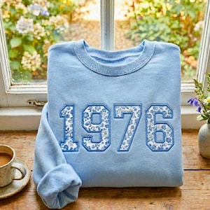 May include: A light blue sweatshirt with the year "1976" embroidered on the front. The numbers are filled with a blue floral pattern and outlined in a darker blue. The sweatshirt is folded on a wooden surface near a window.