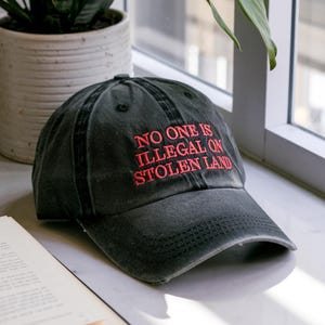 May include: A black baseball cap with the embroidered text "NO ONE IS ILLEGAL ON STOLEN LAND" in red. The cap is made of a worn, distressed fabric and has a curved brim. The hat is sitting on a white surface.