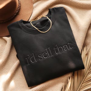K&ouml;nnte beinhalten: Schwarzes Sweatshirt mit dem gestickten Text "I'd sell that". Eine goldene Halskette liegt auf dem Sweatshirt. Das Sweatshirt ist auf einer beigefarbenen Strickdecke gefaltet, mit einem braunen Hut im Hintergrund.