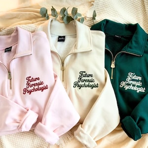 May include: Three quarter-zip sweatshirts in pink, cream, and dark green. Each sweatshirt features the embroidered text "Future Forensic Psychologist" in a contrasting color. The sweatshirts are displayed on a neutral-toned surface.