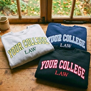 May include: Three crewneck sweatshirts in gray, navy blue, and black. Each features the text "YOUR COLLEGE" in an arched font with "LAW" underneath. The gray sweatshirt has yellow text, the navy blue has white checkered text, and the black has pink text.