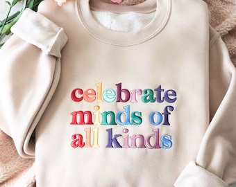 May include: A beige sweatshirt with the embroidered text "celebrate minds of all kinds" in rainbow colors. The sweatshirt has a crew neck and long sleeves with rolled cuffs. The garment is displayed on a soft, textured surface.