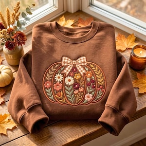 May include: A brown crewneck sweatshirt featuring an embroidered pumpkin design on the front. The pumpkin is decorated with various colorful flowers, leaves, and berries, and is topped with a beige and white gingham patterned bow.