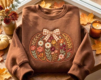 May include: A brown crewneck sweatshirt featuring an embroidered pumpkin design on the front. The pumpkin is decorated with various colourful flowers, leaves, and berries, and is topped with a beige and white gingham patterned bow.