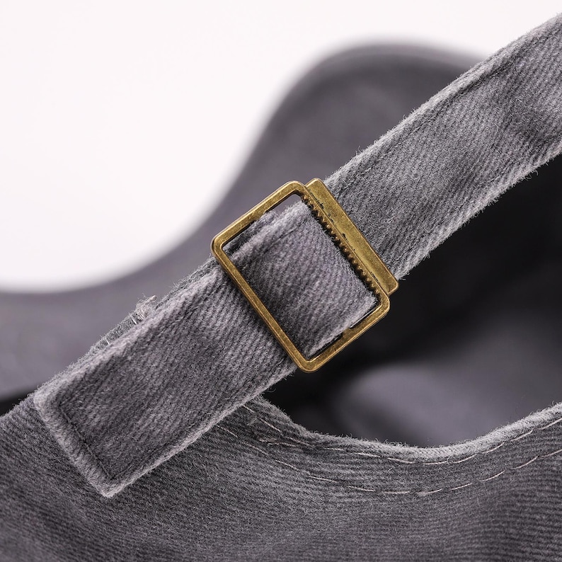 May include: Close-up of a gray baseball cap with a brass-colored metal buckle on the adjustable strap. The cap is made of a textured fabric.