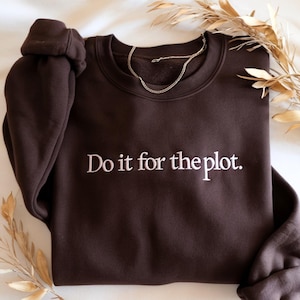 May include: A dark brown sweatshirt with the embroidered phrase "Do it for the plot." A gold chain necklace rests on the sweatshirt. The sweatshirt is folded, with the sleeve bunched up. Dried plants are in the background.