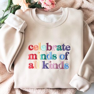 K&ouml;nnte beinhalten: Beiges Sweatshirt mit dem gestickten Text "celebrate minds of all kinds" in Regenbogenfarben. Das Sweatshirt hat einen Rundhalsausschnitt und lange &Auml;rmel mit umgeschlagenen Manschetten. Das Kleidungsst&uuml;ck wird auf einer weichen, strukturierten Oberfl&auml;che pr&auml;sentiert.