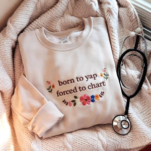 May include: A beige sweatshirt with the text "born to yap forced to chart" embroidered in brown with colorful flowers surrounding the text.
