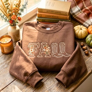 May include: A brown crewneck sweatshirt featuring embroidered text that reads FALL, with the letter F and A decorated with floral motifs and the letters L and L replaced by a pair of embroidered western-style cowboy boots in white thread.