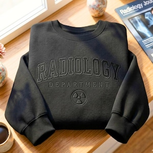 May include: A black crewneck sweatshirt featuring the text RADIOLOGY DEPARTMENT in a dark, embossed-style font across the chest. Below the text is a circular radiation symbol icon. The garment has long sleeves and a ribbed collar and cuffs.