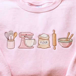 May include: A light pink crewneck sweatshirt featuring an embroidered design of baking tools including a utensil holder with a whisk, spatula, and spoon, a stand mixer, a bag labeled FLOUR, a wooden rolling pin, and a mixing bowl with two spoons.