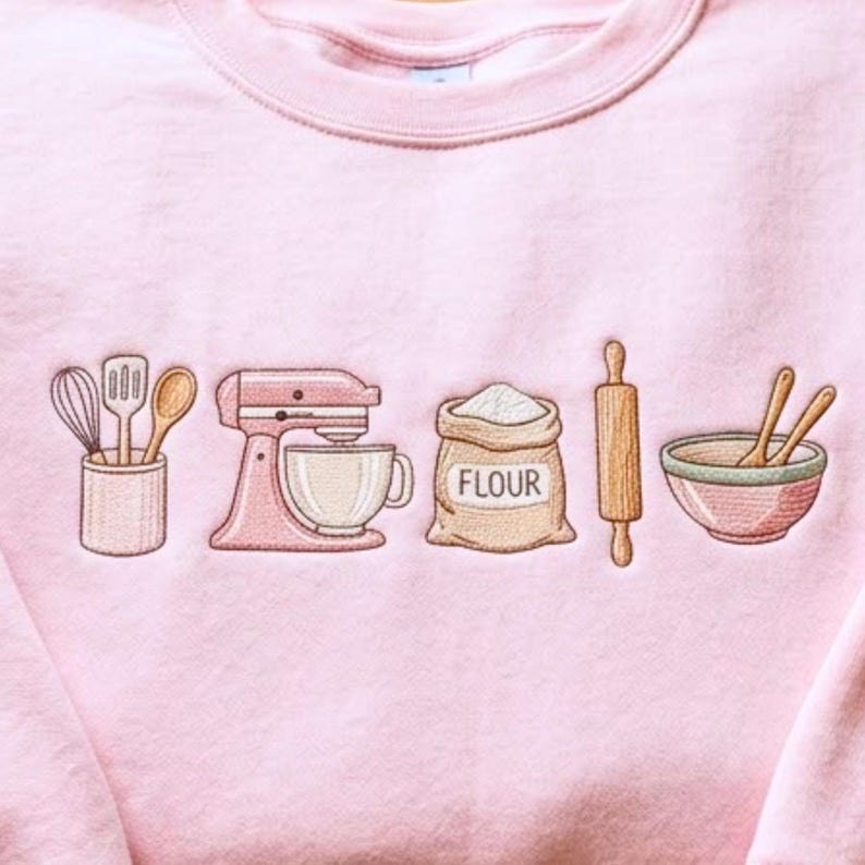 May include: A light pink crewneck sweatshirt featuring an embroidered design of baking tools including a utensil holder with a whisk, spatula, and spoon, a stand mixer, a bag labeled FLOUR, a wooden rolling pin, and a mixing bowl with two spoons.