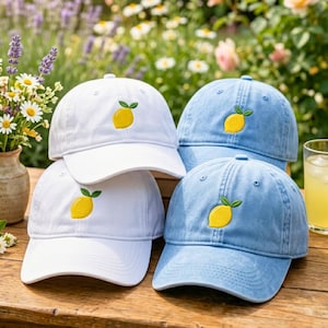 May include: Two white baseball caps and two light blue baseball caps, each featuring an embroidered yellow lemon with green leaves on the front panel. The caps have a structured crown, a curved brim, and a button on the top center of the cap.