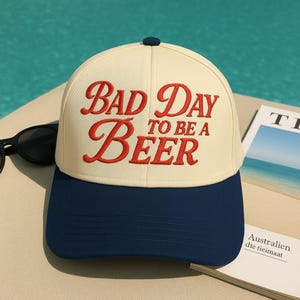 May include: A baseball cap with a cream-colored crown and a navy blue brim. The cap features the phrase "BAD DAY TO BE A BEER" embroidered in red script. A pair of black sunglasses rests beside the cap.